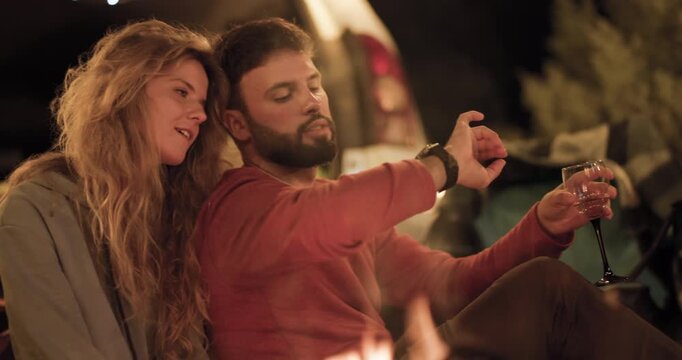 Couple enjoys evening by the campfire while sipping drinks in a cozy outdoor setting under twinkling lights