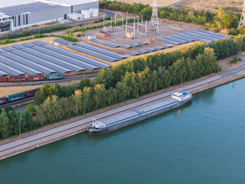 Aerial view of a cargo barge moored on a canal next to an industrial site featuring solar panel arrays, a freight train, and power infrastructure in Genk, Flanders, Belgium.