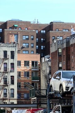 A dense urban landscape with multi-story brick buildings, protruding fire escapes, and a fragment of a car transporter with a white crossover in the foreground, nyc, new york, manhattan
