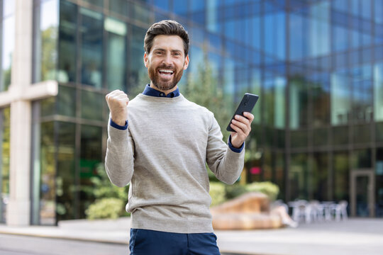 Portrait of a happy young man standing outside, holding a phone, showing a victory gesture with his hand and looking happy at the camera