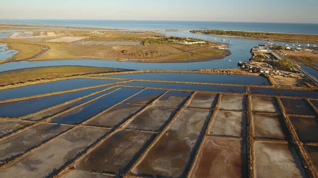 Aerial footage showing the geometric patterns of salt evaporation ponds for sea salt production, with the vast ocean visible on the horizon and a small river flowing towards the coast