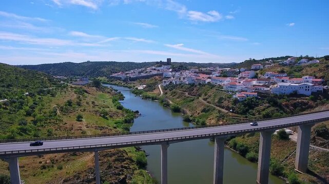 Aerial view showing cars driving on the concrete bridge crossing the guadiana river, with the beautiful and historic town of mertola in the background on a sunny day in alentejo, portugal