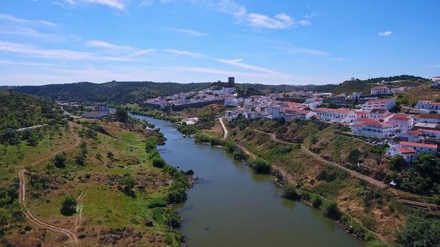 Aerial perspective of the historic town of mertola, featuring its medieval castle and the serene guadiana river flowing through the picturesque alentejo landscape under a bright blue sky