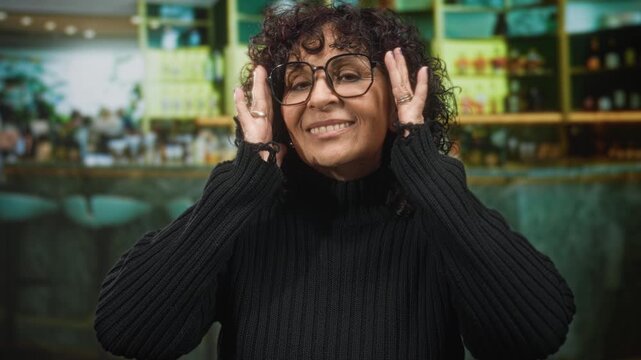 Woman adjusting glasses with hands at temples, smiling in a black turtleneck by a bar counter in studio; joy playfulness.