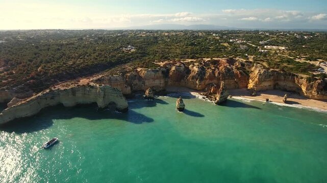 Stunning aerial view flying over the scenic coastline of marinha beach, revealing golden cliffs, natural arches, and a tourist boat cruising on the clear turquoise water of the atlantic ocean