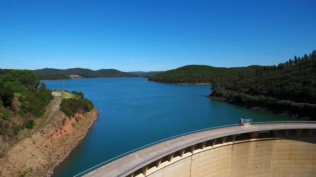 Beautiful aerial view flying backwards, showing the funcho dam, a concrete arch structure, and its vast blue reservoir surrounded by green hills under a clear sky in algarve, portugal