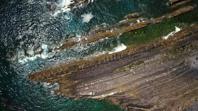 Drone footage capturing the powerful turquoise ocean waves breaking against the unique layered rock formations of a rugged coastline, creating a dramatic and mesmerizing natural display from above