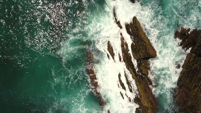 Stunning aerial top down perspective of powerful turquoise ocean waves breaking against a rugged, rocky shoreline, creating white foam and showcasing the raw beauty and force of nature