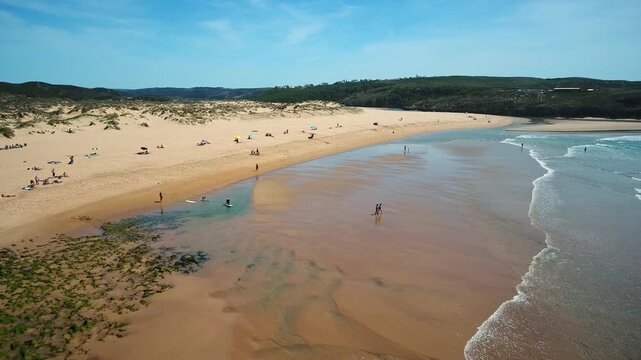 Aerial footage showing the vast sandy shore of amoreira beach in aljezur, portugal, with tourists enjoying the sunny weather and gentle atlantic ocean waves crashing on the coastline