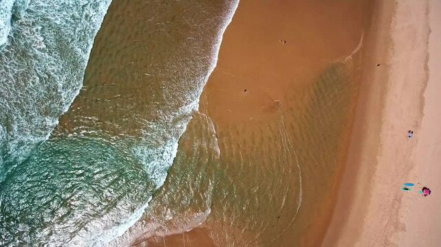 Majestic aerial top down view of turquoise ocean waves gently crashing on a beautiful sandy beach, with people surfing and relaxing on the shore during a peaceful summer day