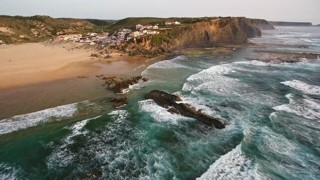 Dramatic aerial perspective of the atlantic ocean waves crashing against the cliffs and sandy beach of praia de monte clerigo, showcasing the charming coastal village in aljezur, portugal
