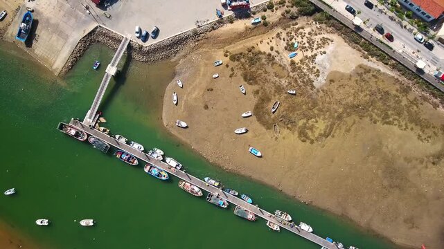 Stunning top down aerial footage capturing a long pier with numerous traditional fishing boats moored in a tranquil green water harbor, contrasting with the sandy, dry tidal flat beside it