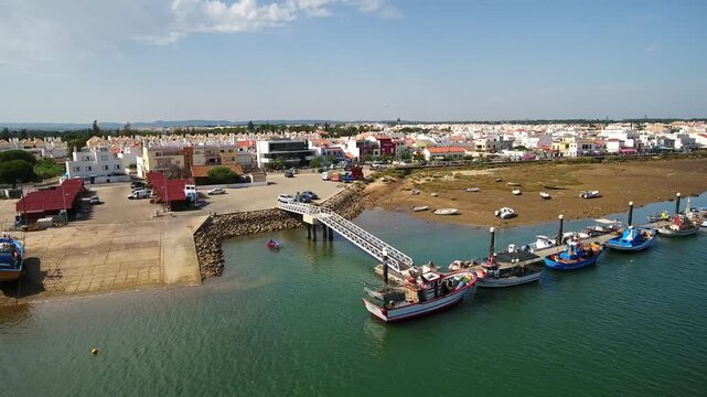 Panoramic aerial view flying over the traditional fishing village of cabanas de tavira, showcasing colorful fishing boats moored at the dock in the ria formosa natural park, algarve