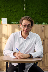 Fototapeta premium Focused young professional writing notes at desk with glasses, white shirt, warm side lighting and green textured backdrop for business planning and productivity concepts