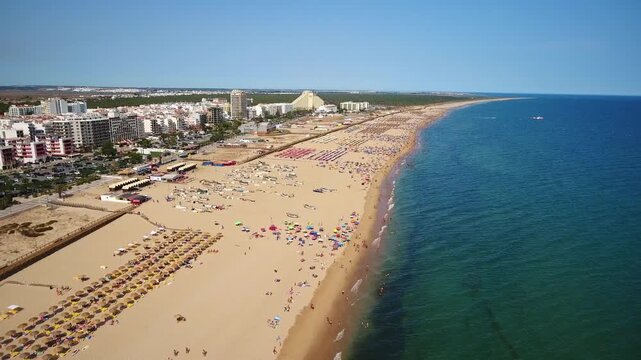 Beautiful aerial view flying along the crowded sandy beach of monte gordo in the algarve, portugal, showing tourists enjoying the summer vacation on a sunny day next to the atlantic ocean