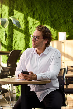 Smiling middle aged man in white shirt sits at round table writing on paper while holding smartphone in bright modern interior with sunlit walls, cabinet and plant