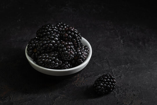Ripe blackberries in white bowl on black background, moody fruit closeup