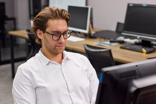 Focused young man working at computer in modern open office, wearing white shirt and glasses, with calm smile, desks and monitors visible in bright workplace, with man, male, office, computer detail