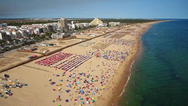 Spectacular aerial perspective flying over monte gordo beach in algarve, portugal, showing a crowded shoreline with colorful umbrellas, tourists sunbathing, and the vibrant blue atlantic ocean