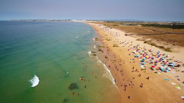 Drone footage capturing the picturesque coastline of gale beach in albufeira, with many tourists enjoying the summer sun, swimming in the ocean, and relaxing under colorful umbrellas