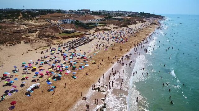 Sunny aerial perspective of gale beach in albufeira, portugal, with crowds of people sunbathing on the golden sand under colorful umbrellas and swimming in the clear turquoise ocean water