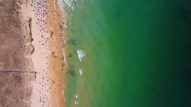 Stunning top down aerial perspective of a crowded sandy beach with colorful umbrellas, people enjoying the sun, and gentle turquoise waves washing ashore on a beautiful summer day