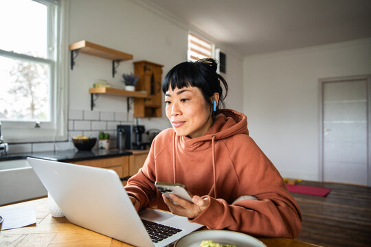 Woman working on laptop with smartphone in home kitchen