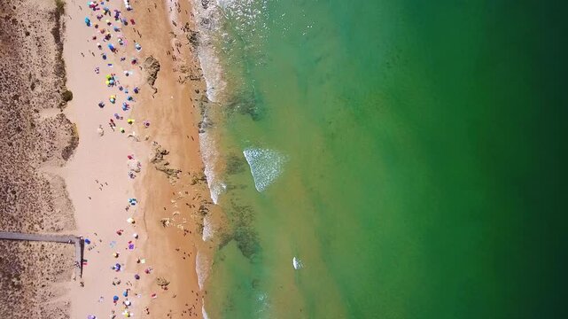 Bird's eye drone footage capturing a beautiful sandy beach coastline with many people and colorful umbrellas, as turquoise ocean waves gently lap the shore on a sunny summer day