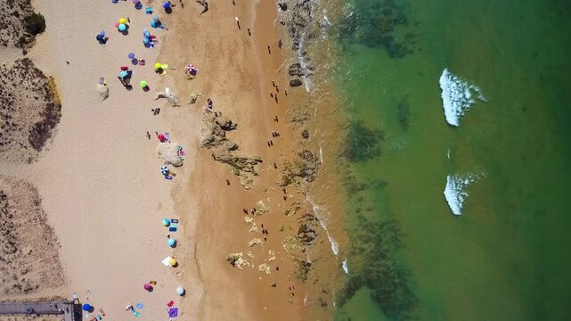 Stunning top down aerial perspective of a crowded beach during a sunny summer day, with colorful umbrellas on the sand and people swimming in the clear green waters of the ocean