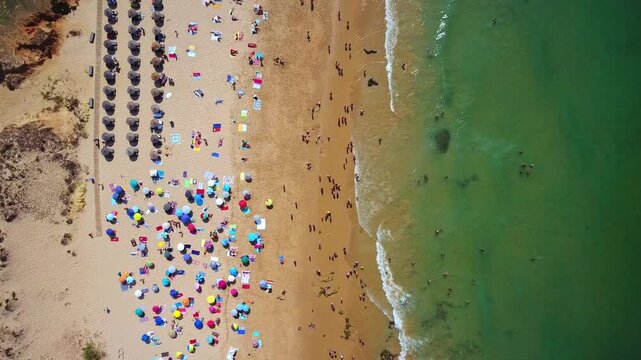 Majestic aerial top down view showing a crowded beach with colorful umbrellas and tourists swimming in the clear ocean water, while the drone flies along the beautiful coastline