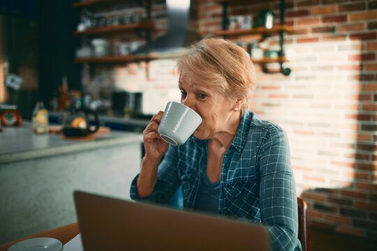 Senior woman drinking coffee while using laptop in home kitchen