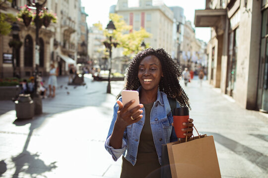 Smiling young woman using smartphone while shopping on city street