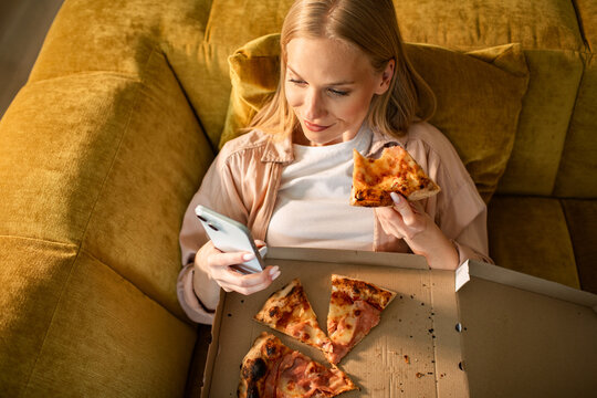 Woman relaxing on sofa eating pizza and using smartphone at home