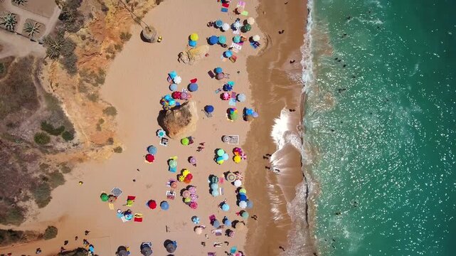Majestic aerial top down view of a crowded beach with colorful umbrellas and people swimming in the turquoise ocean, revealing the sandy coastline and reddish cliffs on a sunny summer day