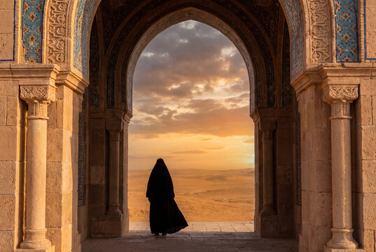 Silhouetted Muslim woman wearing a traditional black abaya looking at the sunset sky through an ornate Islamic mosque archway