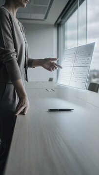 Woman interacts with a translucent digital interface floating above a wooden desk in an office setting. Bright daylight enters through large windows while she points at glowing icons.