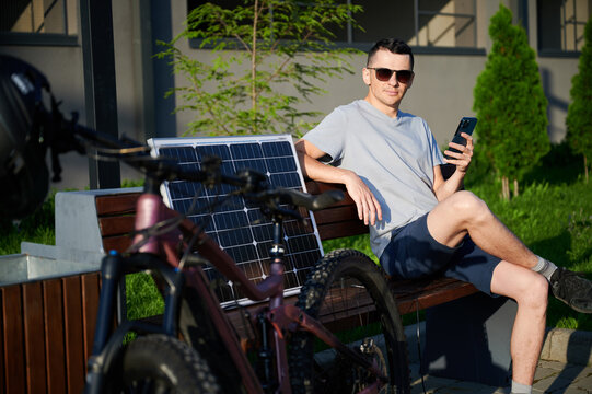 Man cyclist using solar panel for charging electric mountain bike outdoors. Guy in sunglasses sits on bench, holding phone, casually dressed in t-shirt, shorts, enjoying sunny outdoor setting.