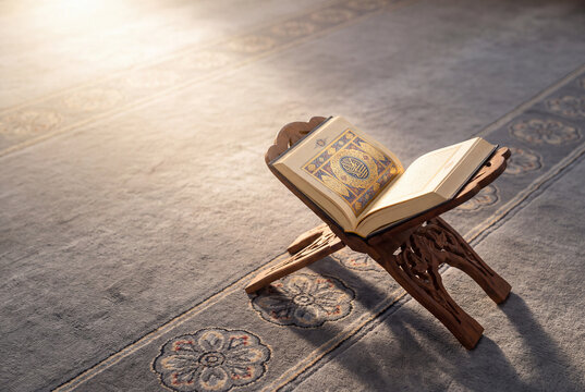 Open holy Quran resting on a wooden stand on a mosque carpet in soft morning sunlight with copy space on a traditional Rihal wooden folding stand