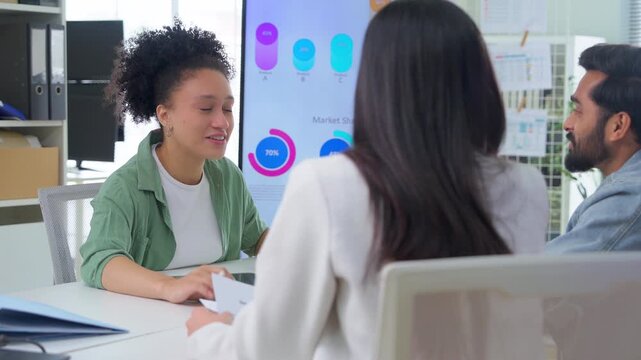 Diverse team of three young professionals discuss ideas around a meeting table with an AI Assistant video presentation playing on a large screen in a modern office.