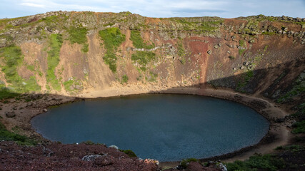 Kerid Crater in Iceland featuring a deep volcanic caldera with a blue crater lake, colorful volcanic slopes, rocky terrain and green vegetation under a clear sky. © Asanee