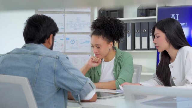 Diverse team of three young professionals discuss ideas around a meeting table with an AI Assistant video presentation playing on a large screen in a modern office.