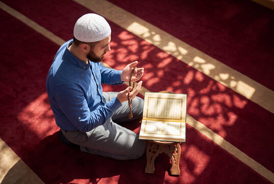 High-angle shot of a young Muslim man praying with prayer beads in front of an open Quran on a mosque carpet