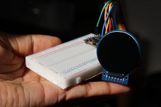 Breadboard connected to microcontroller with jumper cables to round lcd display held in the hand