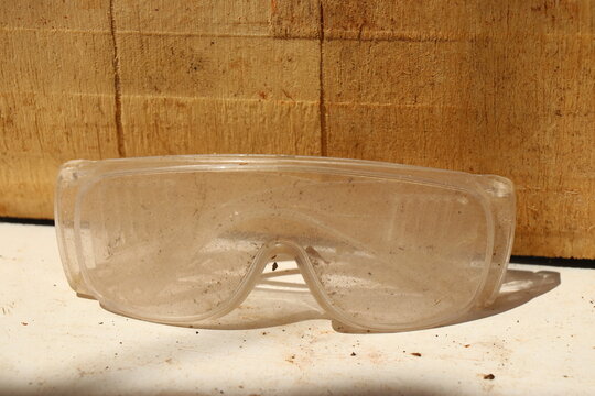 Used clear safety glasses show visible dust and wear isolated against a rustic wooden background