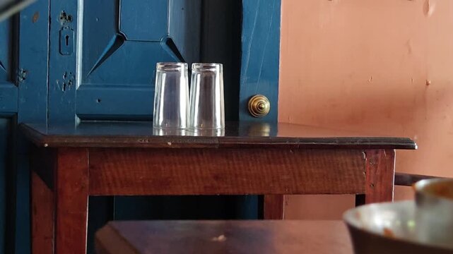 Clean inverted glass tumblers resting symmetrically on a dark wood side table, balancing functional hospitality utility against vibrant architectural panels and quiet restaurant interiors.