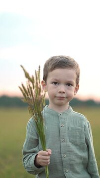Young boy in green shirt holds wild foxtail grass bouquet in soft-focus field at dusk, capturing pure childhood innocence and rural nature connection under pink evening sky.