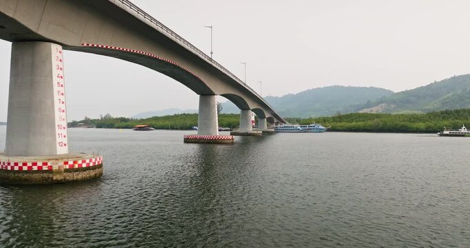 Siri Lanta Bridge spanning calm river in Krabi, ferry boats and dense mangrove forests along distant hilly shores under overcast sky. Travel and explore Thailand infrastructure. Low angle drone flight