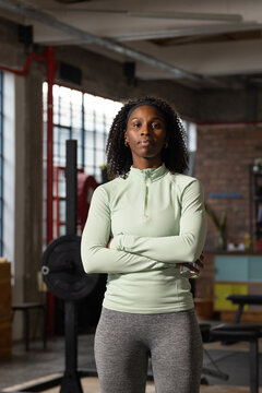 Mid adult woman standing wearing athletic wear with crossed arms in loft gym near squat rack