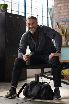 Middle-aged man wearing dark athletic wear sitting on bench in gym holding water bottle smiling