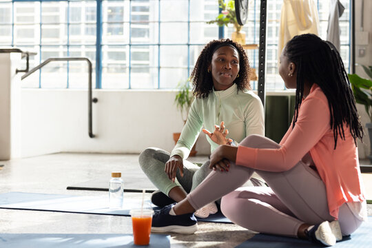 African American female friends in sportswear sitting on yoga mats at studio, holding water bottle
